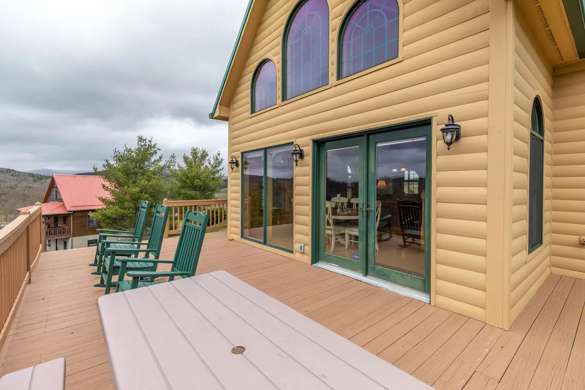 An exterior deck scene showing comfortable seating, a wooden dining table, and expansive views through large windows. The setup is ideal for outdoor dining or social gatherings, with direct access to the house’s interior and an inviting atmosphere for al fresco meals.