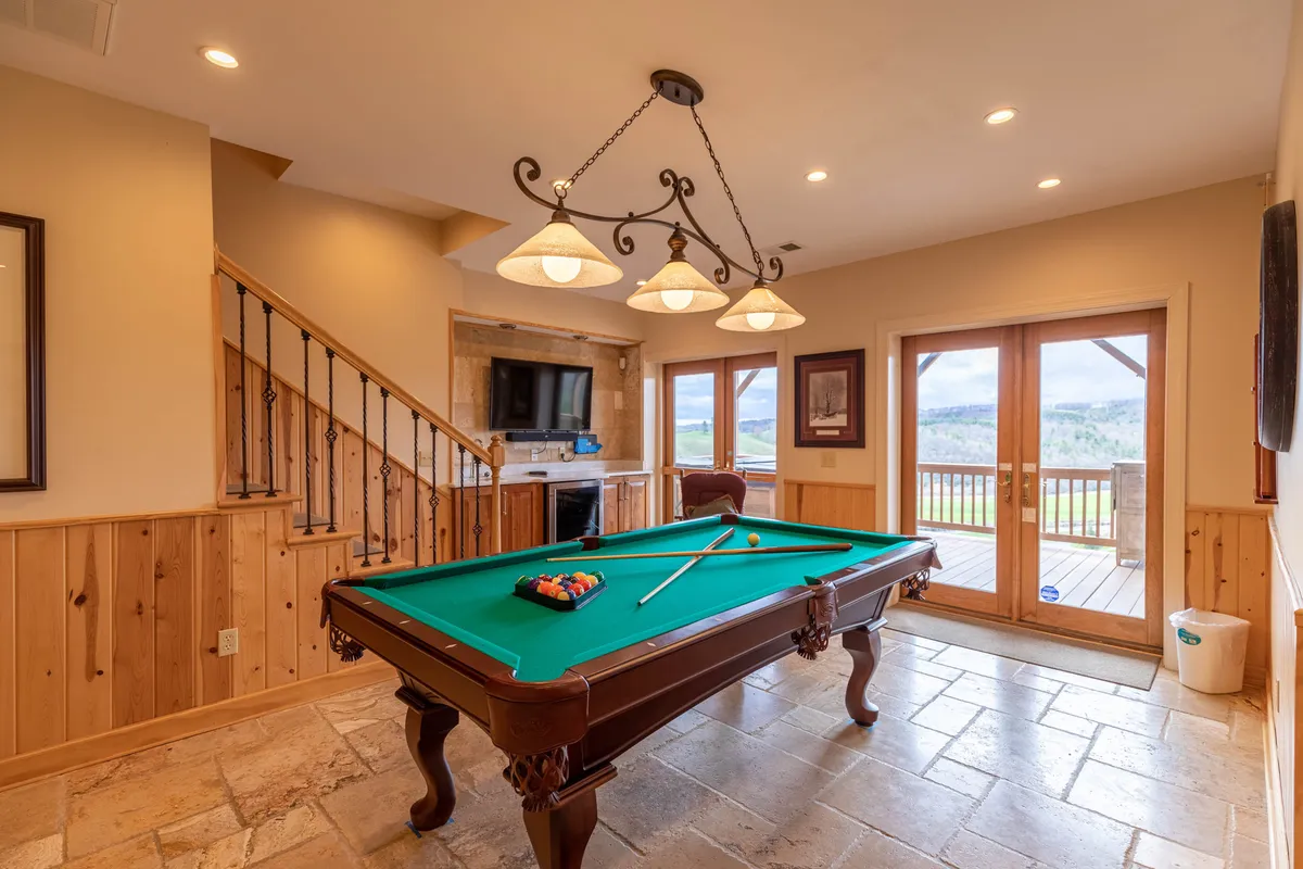 The spacious lower-level game room with a pool table under a warm chandelier is shown, complemented by large windows that offer scenic views. The room feels lively and open, perfect for group activities and indoor entertainment.