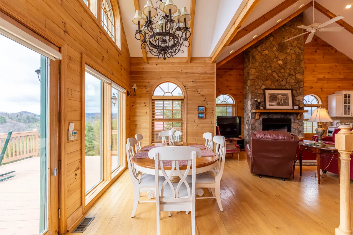 This image captures a warm, sunlit communal space with a large dining table set beneath a stylish chandelier. Expansive windows line the walls, revealing breathtaking mountain and river views. The inviting living area, featuring plush seating and a striking stone fireplace, is perfect for gatherings or quiet relaxation.