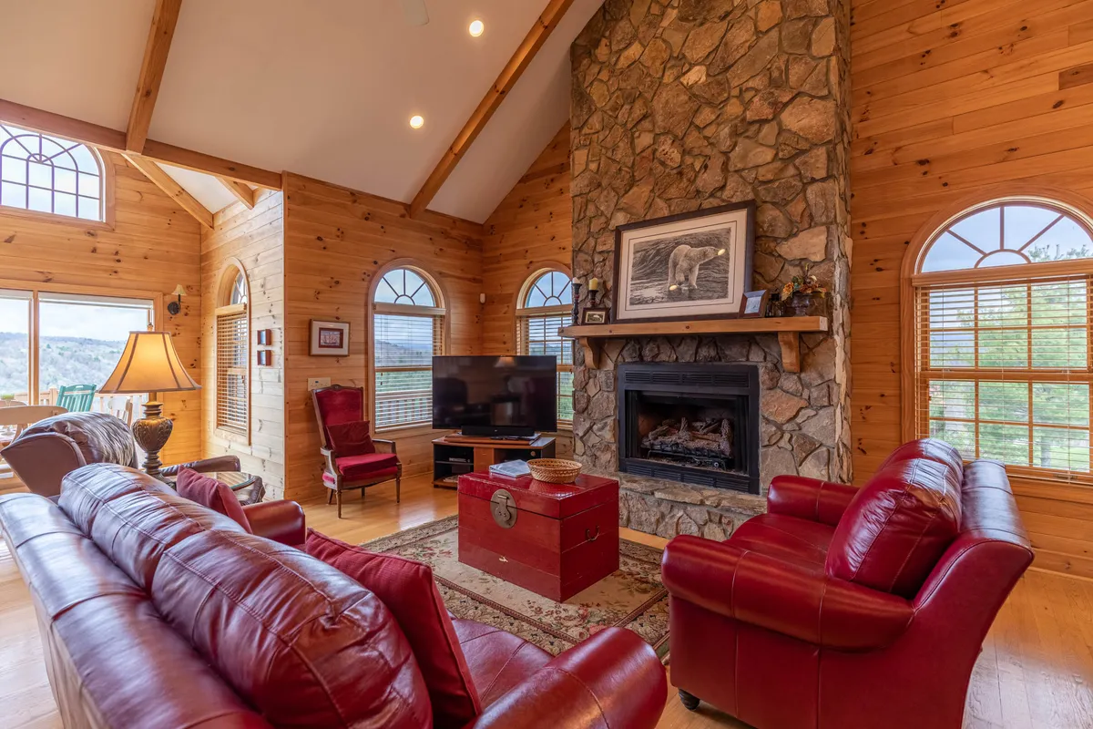 A cozy living room scene with red leather sofas positioned around a grand stone fireplace. Sunlight streams through the large windows, illuminating the plush furnishings and decorative artwork. The view outside hints at the surrounding natural beauty, creating a seamless indoor-outdoor connection.