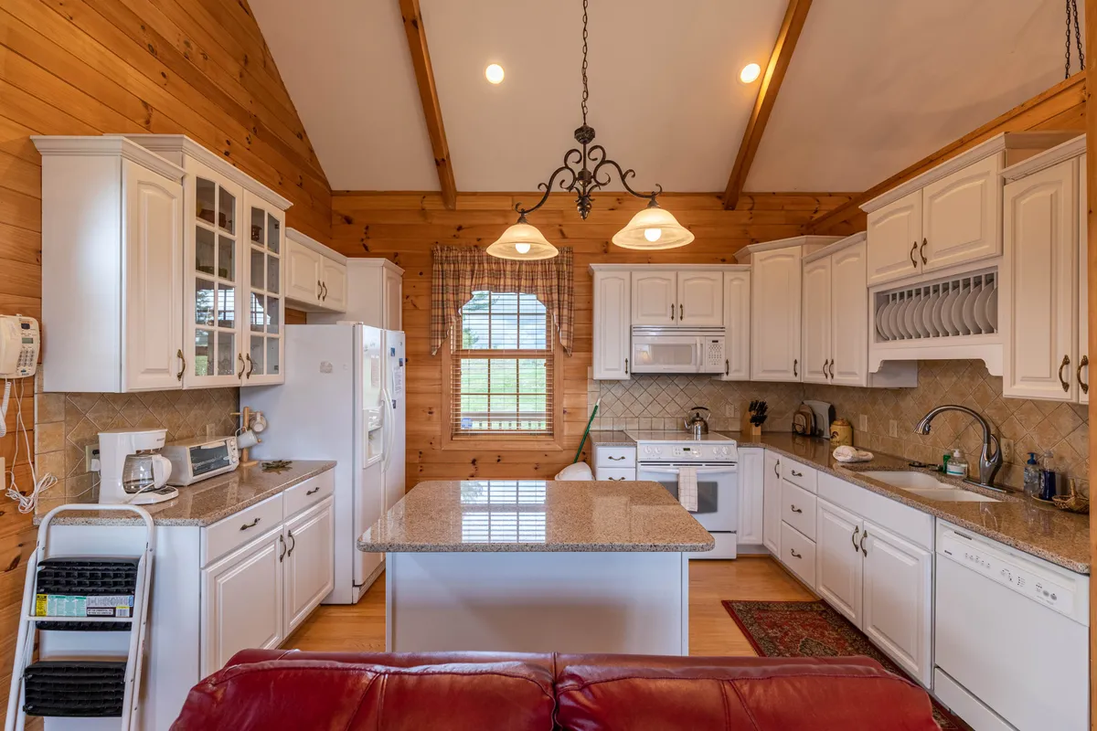 This image features a modern kitchen with a central granite island, white cabinetry, and a large window that brings in ample natural light. The kitchen is well-lit with recessed lighting fixtures and offers plenty of counter space, making it ideal for cooking and entertaining. The overall atmosphere is cozy and welcoming, perfect for gatherings and culinary activities.
