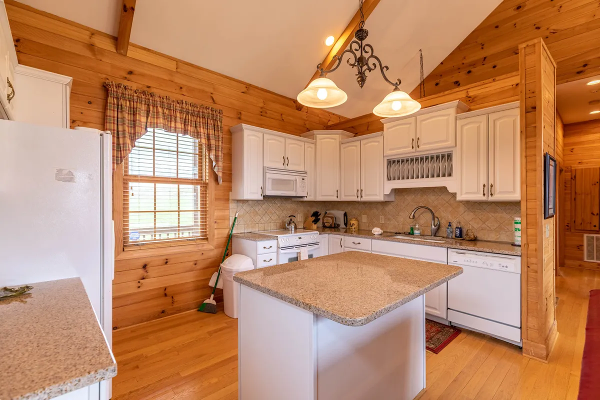 A kitchen scene displaying granite countertops, white cabinetry, exposed wooden beams, and a large window flooding the room with light. The space exudes warmth and is set up for both cooking and gathering, ideal for preparing holiday meals or casual breakfasts.