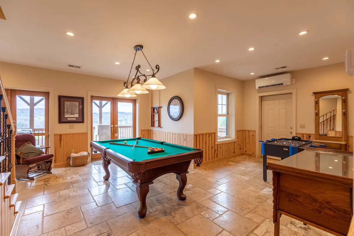 A spacious and inviting game room featuring a pool table and a foosball table, with warm wooden paneling and natural light streaming through the windows. The classic chandelier and comfortable atmosphere set the tone for playful competition and group gatherings.
