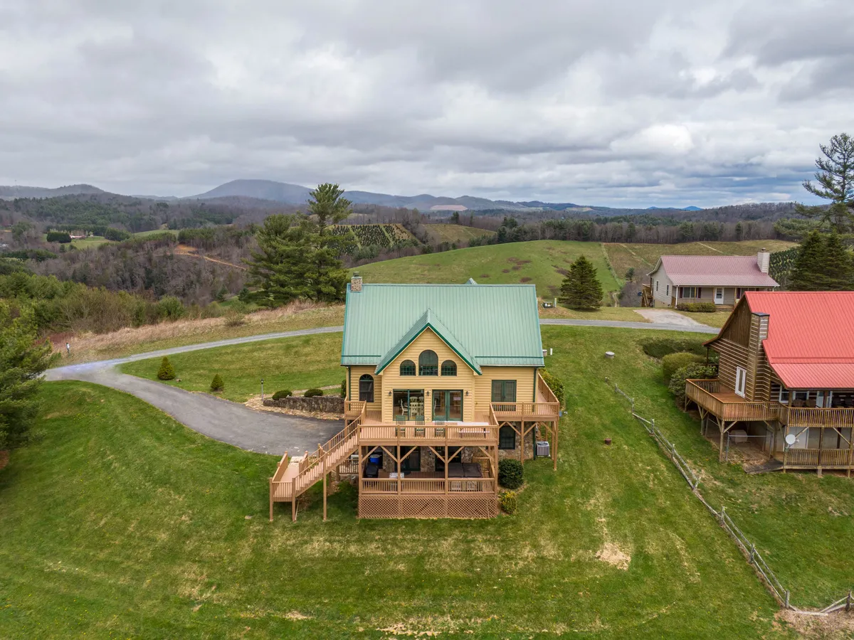 The hero image showcases a charming single-family cabin with a green roof set amid rolling hills and lush mountain scenery. The spacious deck offers inviting seating areas perfect for outdoor gatherings, with expansive views of the natural landscape all around. The tranquil setting promises both privacy and connection to nature, while the deck hints at leisurely mornings, stargazing nights, and easy access to outdoor adventure.