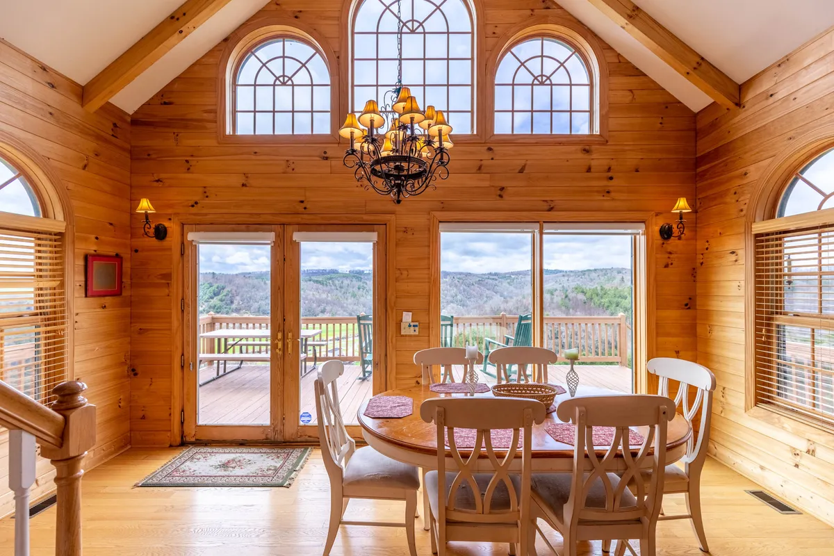 An inviting dining area features a prominent wooden table under a distinctive chandelier. Large windows wrap the space, offering scenic sunrise views and abundant natural light. The wooden walls and décor add a touch of rustic charm, while nearby deck access hints at the close relationship between indoor and outdoor living.