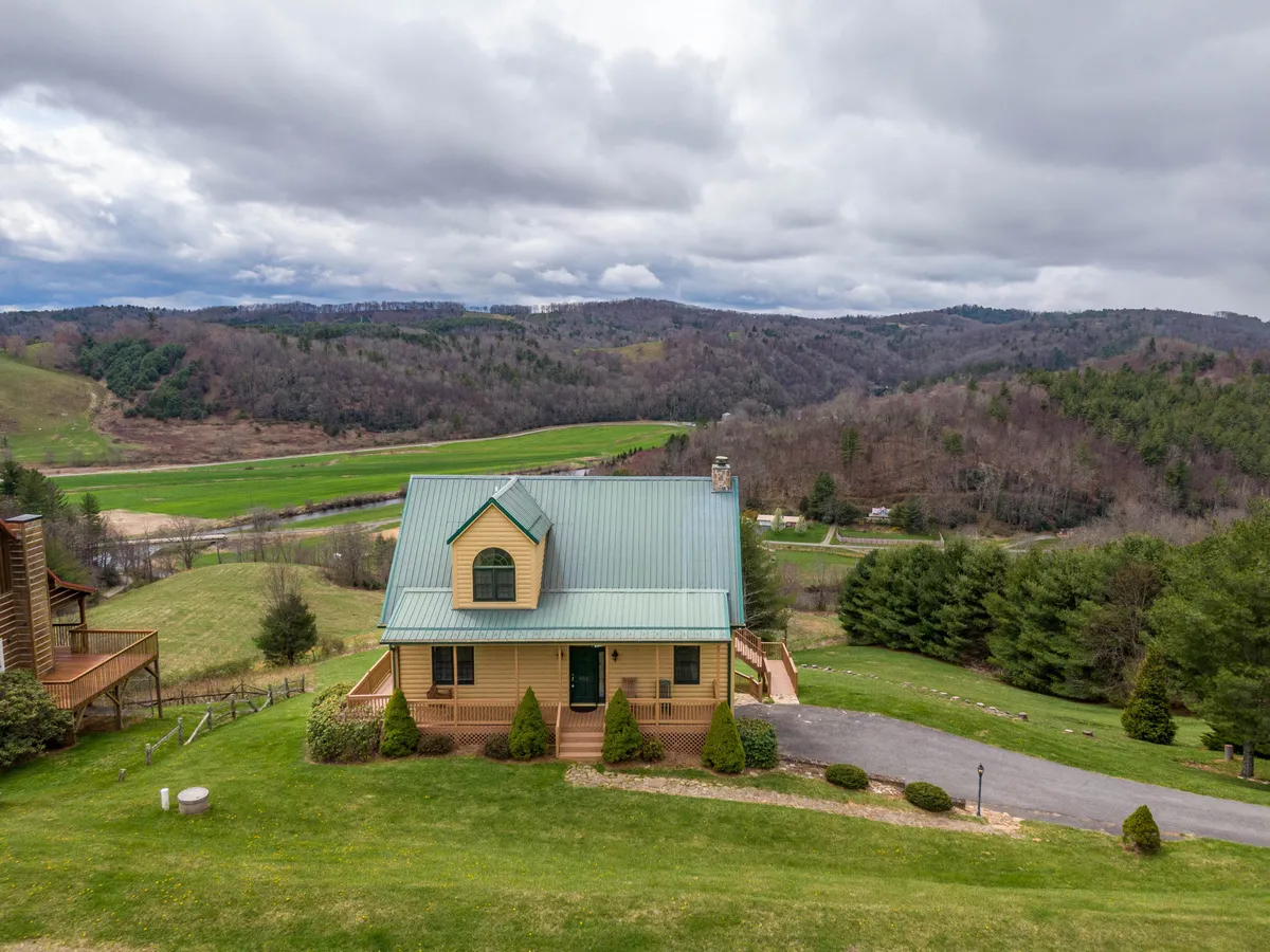 A wide shot of the property showing the house nestled amidst a peaceful landscape with hills and a welcoming driveway. The foreground includes open space suitable for outdoor activities, with mountains in the background enhancing the sense of place.