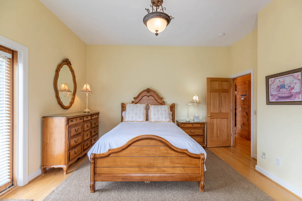 This image shows a serene bedroom featuring a wooden bed with crisp linens, an antique dresser with a mirror, and gentle natural light streaming through the window, creating a restful and welcoming atmosphere ideal for unwinding.